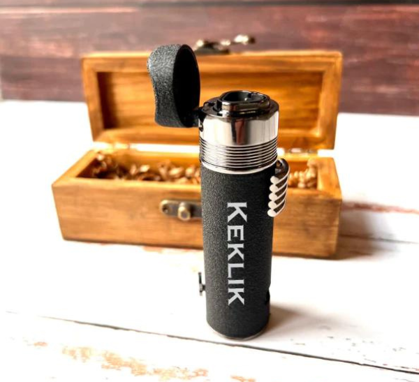 An engraved black torch lighter with its flip top open, standing in front of an open wooden keepsake box on a rustic white plank surface