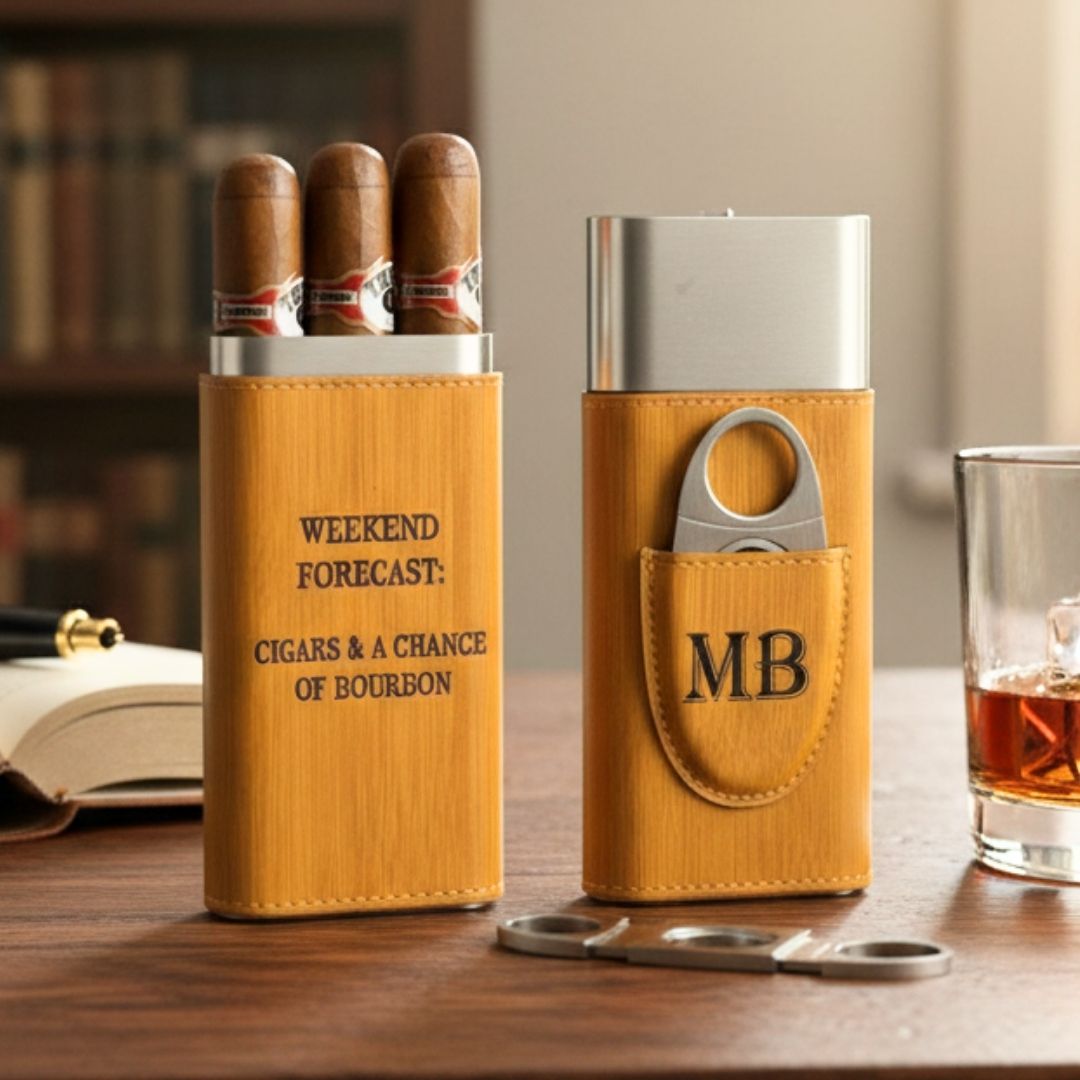 Two tan leather cigar cases with stainless steel tops displayed on wooden desk, left case engraved with sample text "Weekend Forecast: Cigars & a Chance of Bourbon," right case mon