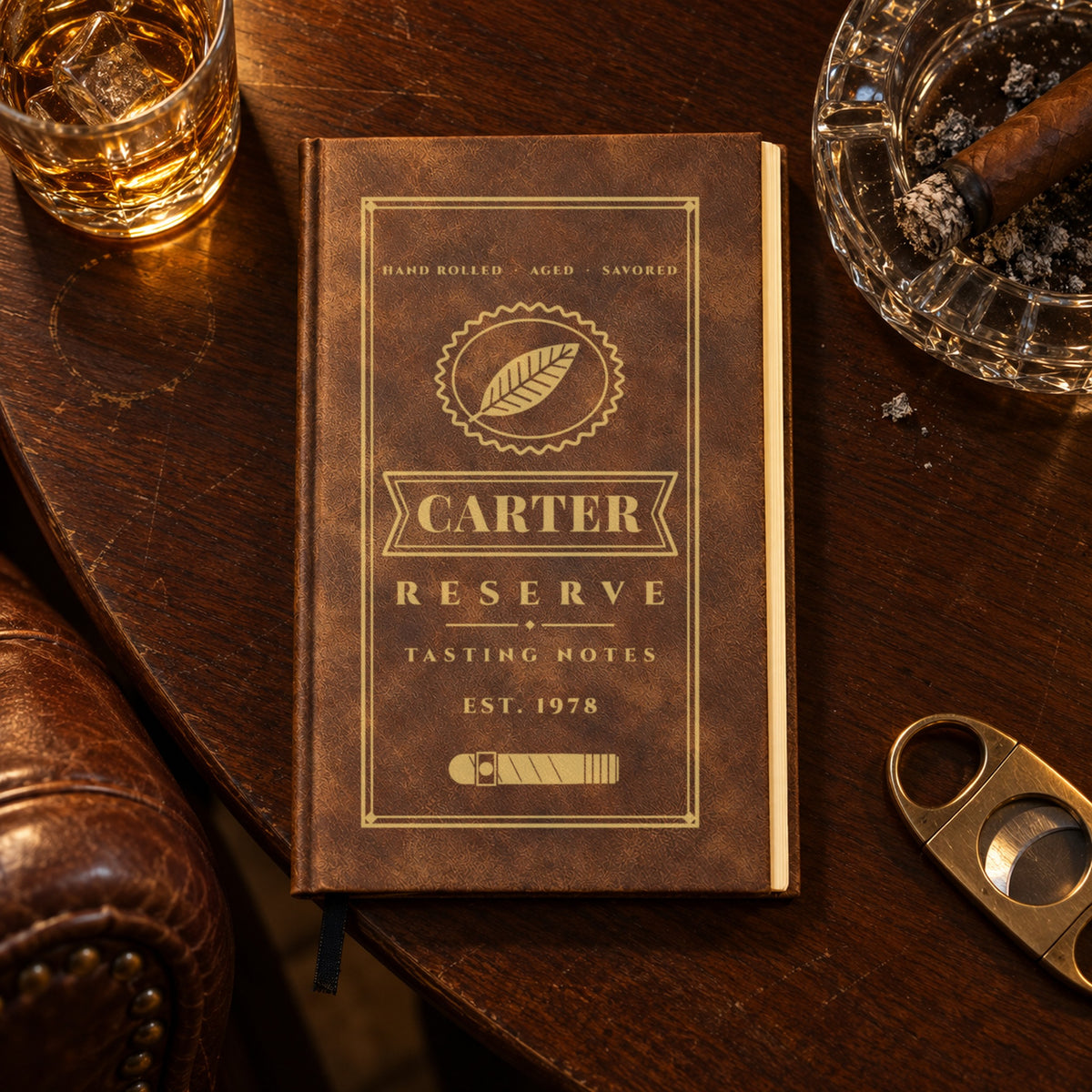 Personalized cigar journal in brown engraved in gold with Carter on a cigar band, Reserve and Tasting Notes, beside an ashtray and a glass of whiskey